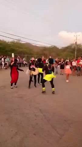 The video shows a group of people performing in an outdoor setting. There are four performers dressed in what appear to be costumes, including black outfits with bright neon yellow and pink accents, and what looks like blackface makeup. One performer is wearing a red dress. They are surrounded by a crowd of spectators standing and watching. The performers seem to be dancing and playing musical instruments that appear to be drums or tambourines. The setting looks like a street or open area in a small town, with houses, foliage, and a hill or small mountain in the background. The video is slightly shaky and appears to be filmed handheld. The overall scene suggests a local festival, carnival, or cultural performance.
