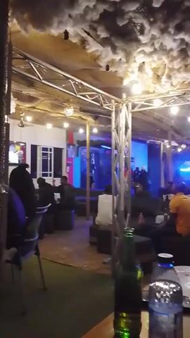 The video shows an indoor lounge area with a casual and slightly dim ambiance. Tables are spread throughout the space, with bottles of beverages (potentially beer and water) visible on some. People are sitting on cushioned seats, engaging in conversations. The ceiling is decorated with a white cloud-like material. Overhead, there are metal beams and strings of lights hanging, adding to the relaxed atmosphere. The sound of a musical performance or amplified conversation can be heard in the background. There is a glass partition in the distance, behind which there appears to be another area with blue lighting, creating depth in the visual layout.