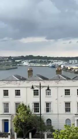 The camera pans right, showing a wide, flat bay or river with a port on its far shore, under a gray, overcast sky. In the foreground are the upper stories of white and tan townhouses with dark slate roofs. A dark lamppost stands near the left corner. The port features warehouses, storage tanks, and a blue ship. The far shore is lined with trees.