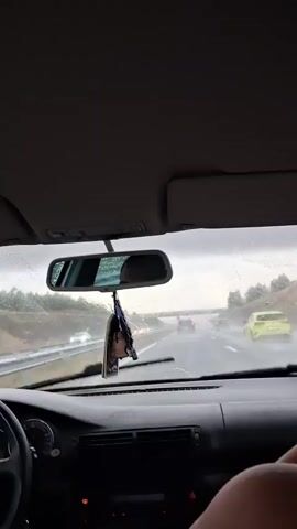 This video shows the interior of a car driving on a highway in the rain. The windshield wipers are visible and active, clearing rain from the view. A car air freshener hangs from the rearview mirror. Other cars are visible on the highway ahead and to the side, some appearing as blurs due to the rain and speed. The overall scene is dim and overcast, indicating poor weather conditions.