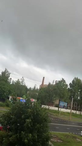 The video shows an overcast sky with grey clouds. In the distance, there is a brick chimney, and some trees. A street with cars is visible in the foreground, and there is a billboard on the side of the road. A bird is flying in the sky. The scene appears to be a somewhat industrial or semi-urban environment.