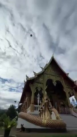 A low-angle shot reveals a temple with intricate gold and green accents. The sky, filled with wispy clouds, forms a backdrop for a small, dark, rectangular object floating near the roof of the temple. A dog lies on the grass at the base of the temple.