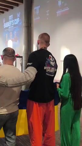 A video captures a scene indoors, possibly an event venue, with a large projection screen displaying text related to "BABAYEV'S CUP". Three individuals are prominently featured in the foreground, with their backs to the camera. On the left, a person wearing a beige shirt and jeans has their arm around the shoulder of the central figure. The central figure, a tall individual, wears a black t-shirt with graphics and text, and orange pants. To the right, a person in a green outfit stands next to the central figure, holding onto a blue and yellow flag, which appears to be the Ukrainian flag. Another person, holding a camera, is visible in the background to the left, near a stage or platform. The environment includes concrete pillars and a wooden ceiling, suggesting an industrial or modern architectural style. Several other people are visible in the background, suggesting a public gathering.