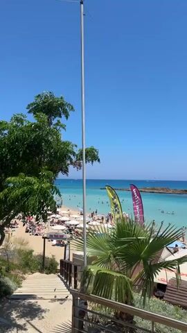 The video shows a crowded beach on a sunny day with clear blue skies. The turquoise water is calm and filled with swimmers and a few boats. People are sunbathing on the sandy beach under umbrellas and lounge chairs. There are also some water sports activities happening in the water, such as jet skiing and paddleboarding. In the foreground, there is a wooden walkway with a railing, and some palm trees and colorful flags. A large red heart sculpture with the words "Love is in the Sea" is visible on the walkway.