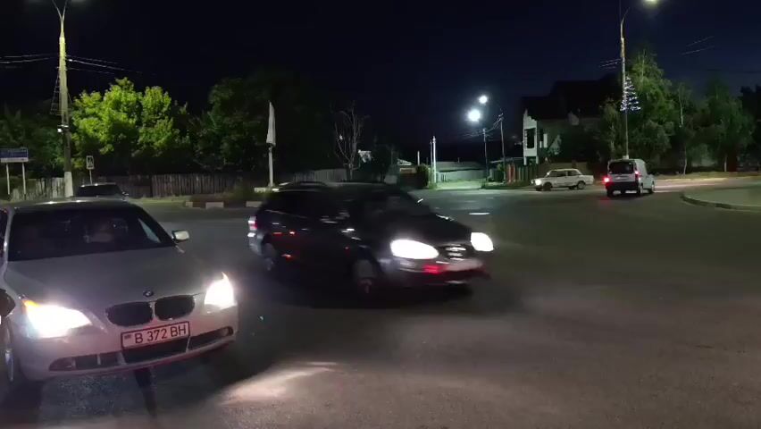 At night, a silver BMW sedan is parked facing forward with its headlights on, its license plate reading B 372 BH. To its right, a dark blue sedan is turning right, its headlights on and its motion captured as a blur. In the background, trees and streetlights are visible, along with houses and other vehicles on the road.