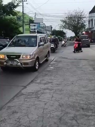 A gold SUV is parked on the left side of a paved road, with its front passenger door open. Several motorcycles are riding in a procession down the right lane of the road. The sky is overcast. Buildings and trees line the left side of the road. There are power lines overhead. The motorcycles are moving away from the camera, and their occupants are facing forward. The lead motorcycle is red, and it is followed by several dark-colored motorcycles. A dark-colored SUV is visible in the distance on the right side of the road.