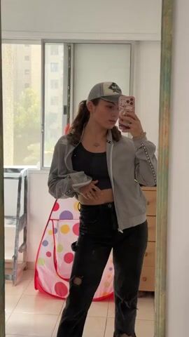 A woman is taking a selfie in a mirror. She is wearing a gray jacket, a black crop top, black jeans with rips, and a baseball cap. Her hair is curly and pulled back. She has rings on her fingers and a bracelet on her wrist. The phone has a pink floral case. The room has a window overlooking a city, a dresser, a baby play tent, and a small ladder. The mirror has a green and brown frame. She poses in two positions: facing forward and then twisting to the side to show her back.