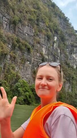 The video shows a woman in an orange life jacket smiling and waving into the camera. Her sunglasses are perched on her head.  Behind her is a large rocky cliff covered in trees.  There is a river on the left side of the frame. The sky is overcast. The woman says "We are in Vietnam."