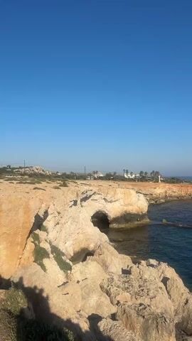 A video shows a rocky coastline with clear blue water. In the first scene, a cave is visible in the rock face. The second scene pans out to show more of the coastline and the vast ocean. The third scene focuses on a large rock formation rising out of the water. The fourth scene shows several people gathered on a rocky outcrop near the water's edge. The final scene features a natural rock bridge spanning the water, with a woman standing on top of it and other people visible on the rocks below. The sky is clear and blue throughout the video.