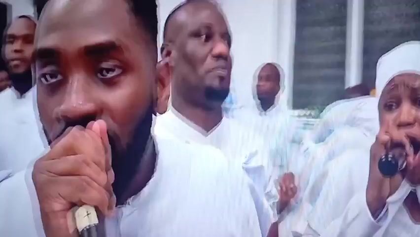 The video shows a group of people, mostly men and women, dressed in white robes and head coverings, gathered in what appears to be a religious or ceremonial setting. The camera focuses on several individuals, including a man in the foreground who is coughing or touching his face, and a woman holding a microphone. The lighting is dim, and the scene is somewhat blurry due to movement and image processing. Multiple overlapping images of people suggest a fast-paced or disorienting visual effect. The overall impression is that of a spiritual or emotional gathering, possibly a church service or a similar event.