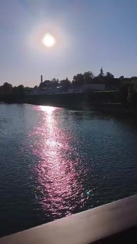 The video shows a panoramic view of a river on a sunny day. The sun is high in the sky, casting a bright reflection on the water's surface. On the left bank, there are buildings, a dock with boats, and trees. The right bank is lined with trees and appears to be a more natural landscape. There are bridges visible in the distance. The camera pans slowly to the right, offering a wider view of the river and its surroundings.