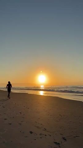 A lone female figure is captured in silhouette as she runs along a sandy beach. The sun is setting or rising, casting a warm golden glow across the scene. Gentle waves lap at the shore, and the sky is a clear, pale blue transitioning to orange near the horizon. The runner's shadow is elongated and stretched across the sand, emphasizing the low angle of the sun. The overall atmosphere is serene and tranquil.