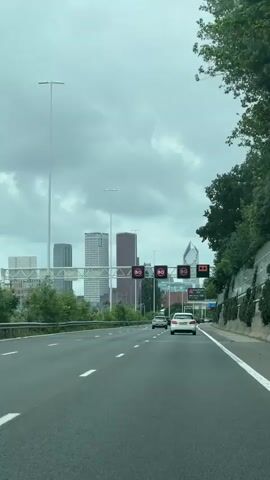 The video shows a view from the passenger seat of a car driving on a highway. The sky is overcast. In the distance, several modern skyscrapers are visible. Above the highway, a series of electronic signs are mounted on a gantry. Three of these signs display a red circle with the number 80 inside, indicating a speed limit of 80 kilometers per hour. The fourth sign to the right displays an orange arrow pointing to the right, suggesting an upcoming lane closure or change in direction. Several cars are driving on the highway ahead of the camera car. The highway has multiple lanes, and white lane markings are visible. Lush green trees and vegetation line the right side of the highway.