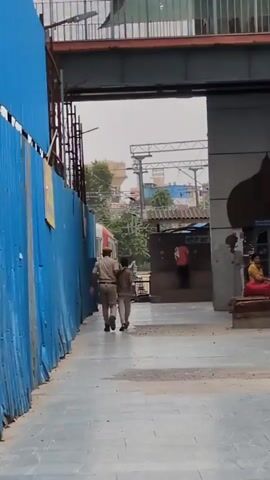 A police officer escorts a young person along a platform at a train station. The officer has his arm around the young person's shoulders. Both are walking away from the camera, towards a train on the left side of the frame. A blue metal fence is to the left of the officer and the young person. In the background, there are train tracks and overhead power lines. The scene appears to be outdoors during the daytime.