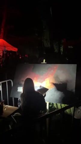 The video shows a person painting a large canvas in an outdoor, nighttime setting. The canvas depicts abstract, fiery imagery with vibrant reds and oranges against a dark background. The artist, silhouetted from behind, is seated and appears to be focused on their work. A bright light illuminates the canvas and the artist's immediate workspace. In the background, a red tent is visible, with the silhouettes of other people standing around it. The overall atmosphere is dark, with artificial lighting playing a significant role in highlighting the artistic activity.