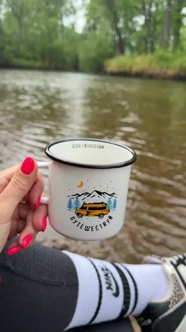 A person wearing black athletic pants, white socks, and grey New Balance sneakers is sitting on a rocky bank next to a river, holding a white camping mug in their left hand. The mug has a black rim and handle. It features a design of a yellow van beneath snow-capped mountains, pine trees, and a crescent moon, with words in Russian printed above and below the image. The person's fingernails are painted bright pink. The river water is murky, and a green, forested bank is visible in the background.