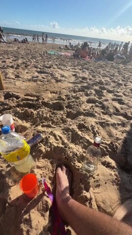 The video captures a sunny beach scene with a cluttered foreground revealing signs of revelry. An individual's foot rests near empty bottles of Black Elephant vodka and juice, a crumpled Red Bull can, and a plastic cup. As the camera pans upward, a vibrant beach unfolds, populated by numerous individuals. Some are sunbathing on towels, while others stroll along the shoreline, dipping their feet in the water. The ocean stretches to the horizon, punctuated by gentle waves under a clear blue sky adorned with scattered clouds. The scene suggests a relaxed, communal atmosphere typical of a day at the beach.