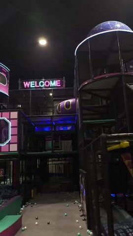 The video shows an indoor playground with neon lights and a futuristic theme. The playground features multiple levels of climbing structures, slides, and a ball pit. The walls and decorations are brightly colored with pink, purple, and blue neon accents. Signs that say "WELCOME" and "HELLO" are visible. Several people, including children and adults, are present in the playground, some are sitting and looking at their phones, others are walking or playing in the ball pit. The lighting is dim, with spotlights highlighting different areas of the playground. The overall atmosphere appears to be playful and engaging, designed for entertainment.