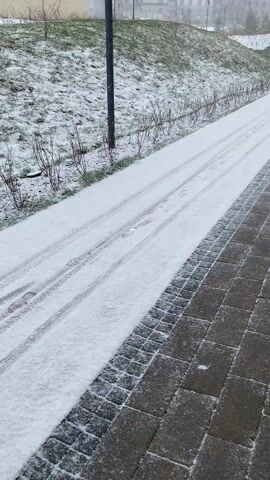 The scene is a slightly elevated sidewalk on a snowy day. The sidewalk is made of gray pavers, and snow covers a strip of grass and bare bushes to the left of the paved path. Tyre tracks are visible in the snow. In the distance, apartment buildings and streetlights are seen through the falling snow. The overcast sky suggests a cold, wintry atmosphere. The camera remains static throughout the video.