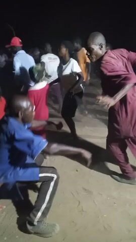 A crowd of people dance in a dirt lot at night. The people are dressed casually. One man in a blue shirt and track pants is facing the camera and dancing, as is a man in a red suit. A young girl in a red dress and a woman in a white shirt and black skirt are also dancing. Other people are milling around in the background. The scene is lit by artificial light, and the quality of the video is poor.