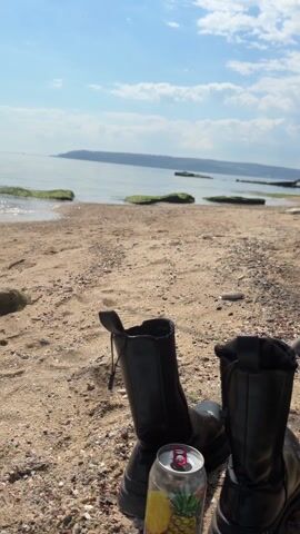 The video shows a beach scene. Initially, a pair of black boots and an open beverage can sit on the sandy beach. The camera pans upwards, revealing a view of the water with some mossy rocks close to the shore. Further out, a landmass is visible in the distance. The camera continues to pan across the water, and a large ship comes into view. The sky is blue with some clouds.