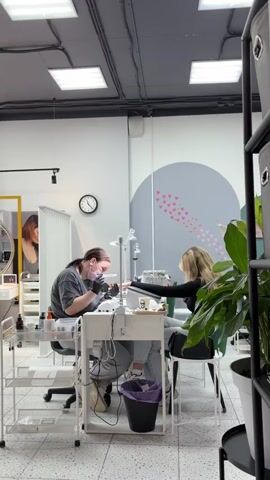 The video shows a nail salon with two women. One woman, wearing a grey shirt and a pink mask, is working on the nails of the other woman. The woman receiving the service has blonde hair and is wearing a black top. Both women are sitting at white tables, each with a lamp. There are various nail care products and equipment visible. The salon appears to be modern and clean, with white walls and a grey accent wall with pink hearts. A clock is visible on the wall, showing approximately 8:12. There is also a plant in a pot in the foreground.