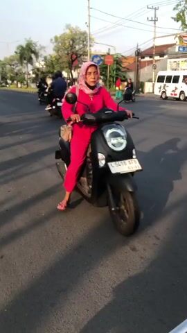A woman wearing a pink hijab and a pink tracksuit rides a black scooter. She is looking at the camera. The scooter has a license plate that reads "B 6498 AFB". She is on a paved road with a white line marking the lane. There are other scooters and motorcycles in the background. The environment appears to be a city street with buildings and trees visible. The sun is shining, casting shadows on the road.