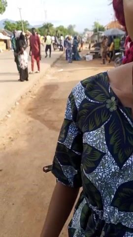 The video begins with a close-up of a woman speaking to the camera. She has dark skin, dark eyes, and her hair is styled with red extensions. She is wearing a blue and green patterned dress with a dark necklace that has a pendant. In the background, a street scene unfolds with several people walking and standing. Some individuals are dressed in traditional African attire. The environment appears to be outdoors, with a dirt road or pathway beside a paved road. The lighting suggests it is daytime. The woman appears to be animated as she speaks, with her mouth open. The video then cuts to a wider shot of the street with the woman still visible in the foreground. Other people are walking in both directions on the street and sidewalk. The setting seems to be a town or village, with buildings visible in the distance. The overall atmosphere suggests a candid street view or possibly a gathering.
