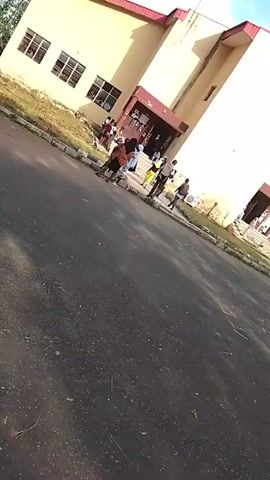 The video shows a low-angle shot of a paved road leading towards a two-story building. A group of people are standing near the building's entrance. The building is painted in light yellow and has a red-brown roof. It has windows and doors, and appears to be an academic building. The road is paved, and there is a small grassy area between the road and the building. The camera is held at a tilted angle, making the scene appear slightly off-balance. The sky is visible in the background.