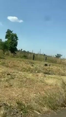 The video shows a white ambulance with a flat tire parked on the side of a road with its back door open. A man in a uniform stands near the road. Further down the road, a small group of people are standing together, some dressed in suits. The setting appears to be a rural area with dry grass and scattered trees under a clear blue sky. The video is taken from inside a passing car.