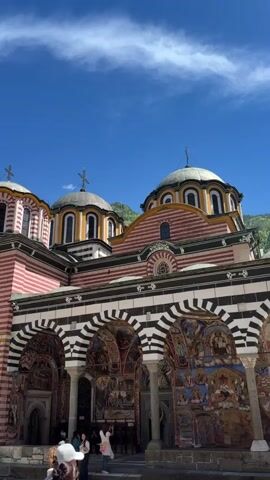 The video shows a low-angle view of the Rila Monastery in Bulgaria. The camera slowly pans upwards to capture the architecture of the building. The building features red and white striped walls, domes with gold accents, and a colonnaded portico adorned with colorful murals. People are seen standing and walking near the entrance of the building. The sky above is clear and blue with some wispy clouds. Lush green trees are visible in the background.