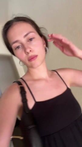 The video shows a young woman, likely in her late teens or early twenties, standing in a hallway. She's wearing a black spaghetti-strap dress and has a black shoulder bag with a gold buckle. The woman has fair skin, light brown hair pulled back from her face, and is wearing silver hoop earrings. She is looking directly at the camera, and she runs her hand through her hair. She appears to be speaking, although the audio is not available. The background shows a beige wall and a marble floor. The lighting appears to be indoor, likely fluorescent. The camera seems to be held at a slightly high angle, suggesting a selfie perspective.