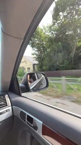The video shows a view from inside a car driving down a road. In the first clip, a side mirror is visible, reflecting a person in the passenger seat. The second clip shows the road ahead with other vehicles, buildings, and trees lining the sides. The sky is blue with some clouds. The third and fourth clips continue to show the view from the car as it drives along the road, with more buildings and vehicles visible.