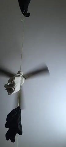 A ceiling fan with its blades spinning rapidly is shown. A string is attached to the center of the fan and hangs down. On the string, near the fan, a white glove is attached, appearing to be suspended from the string. Below the glove, another dark colored object, possibly clothing, is hanging from the string. The fan is mounted on a ceiling, and the background is a plain, light-colored wall. The lighting suggests an indoor environment.