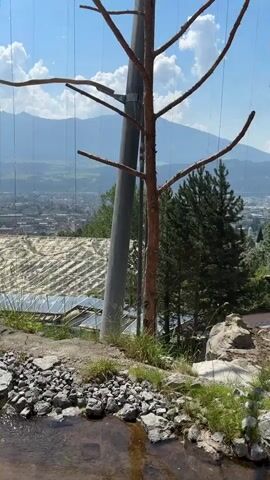 The video shows a view from a rocky, elevated area overlooking a city and mountains. In the foreground, there is a small stream flowing over rocks. A large metal pole and some dead branches are prominent. Further in the background, a large structure with solar panels is visible, along with trees and the city. The video then pans to show an enclosure with a mesh roof, containing rocks, trees, and two people walking on a paved path. Another person is seen standing near a rock formation within the enclosure. A bird of prey is perched on a branch inside the enclosure. The sky is blue with some white clouds.