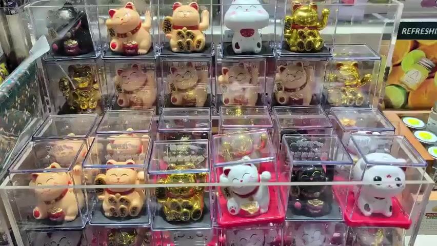 This media file displays a collection of numerous "maneki-neko" or "beckoning cat" figurines, presented in clear plastic display boxes. The figurines are arranged in tiered rows, creating a visually dense display. They vary in color, including gold, white, black, and a peachy-pink hue. Many of the cats have their paws raised, a characteristic gesture of maneki-neko, which are often believed to bring good luck. Some figurines are adorned with small gold coins or other decorative elements.

The background shows elements that suggest a retail or market setting. To the right, a sign with the word "REFRESHING" is visible, along with images of what appear to be fruits or preserved goods in jars, such as limes and peaches. This suggests the items are for sale, possibly in a gift shop, souvenir store, or a market stall in Thailand, given the location context.

The lighting appears to be bright and artificial, typical of an indoor retail environment, and there are no clear indications of the time of day or weather. The focus is entirely on the product display, with no people visible in the immediate foreground or on the display itself. The overall emotion conveyed is one of abundance and a focus on decorative, possibly lucky, items. The image is static, with no discernible activity taking place beyond the presentation of the objects.