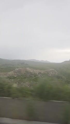 The video shows a landscape from a moving vehicle. The foreground is blurred green foliage and a gray stripe, indicating it is a side view from a car or bus. The midground is a rolling green hill with some rocky outcrops. The background is a hazy expanse of hills under an overcast sky. There are no people or distinct objects visible.