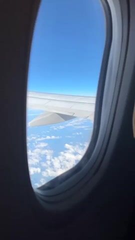The video shows a view from an airplane window. The sky is a clear, bright blue. Below the airplane, a layer of white, fluffy clouds stretches across the landscape. The wing of the airplane is visible in the upper portion of the frame, extending out from the side of the aircraft. The wing is white and appears to be in good condition. The sun is shining, casting light on the clouds and the wing. The overall impression is one of a peaceful and serene flight.