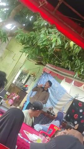 A young man in a brown jacket holds a microphone and sings into it. He stands under a red and white striped awning, in front of a blue tarp with a floral pattern. He is in an outdoor setting, with green foliage visible overhead. A group of people are seated in red chairs at tables covered with red tablecloths, some of whom appear to be watching and listening to the singer. The overall atmosphere suggests a casual gathering or event.