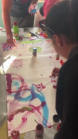 A woman in sunglasses is painting on a large sheet of paper. The paper is covered in abstract designs, flowers, and a ballerina sketch. There are other people gathered around the table, and various art supplies are scattered around, including paint bottles, brushes, and containers of glitter. The scene appears to be a collaborative art activity.