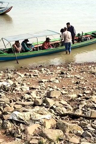 A long green boat with a blue stripe and a canvas roof is docked at a rocky shore. Four people are seated inside the boat, and two people stand at the stern. One of the standing individuals is partially submerged in the water, reaching into the boat. In the background, another boat is visible on the water, and the environment appears to be a body of water with a shoreline covered in rocks and debris.