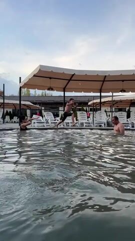 A woman standing in a pool of water throws water on a man who is swimming. The man appears to be surprised, though he is laughing. The scene is outdoors. There are lawn chairs and large umbrellas alongside the pool. It appears to be a spa resort.