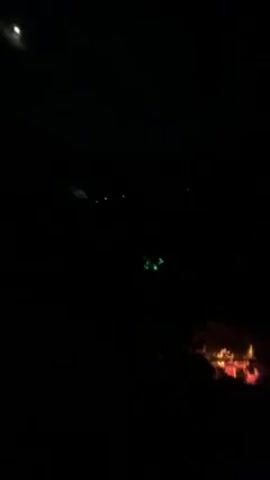 The video takes place at night, in an outdoor setting. Initially, a campfire is visible in the foreground, and flashing colored lights appear in the distance. The background then shows a large area illuminated with red light, possibly from another fire. Finally, the scene shifts to a group of people standing in front of a cliff face lit with green light. There is also a spotlight illuminating the scene.