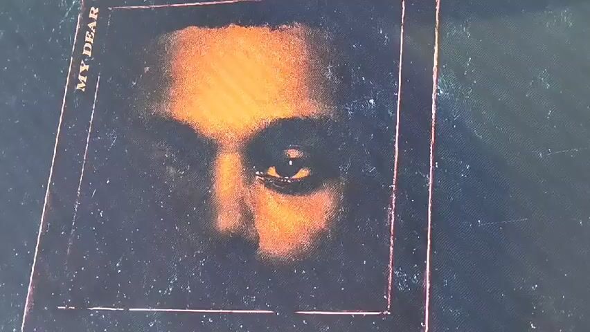 This media file is a close-up shot of a framed artwork, likely a album cover or print. The artwork depicts a person's eyes and forehead in a shadowy, distressed style with warm, orange-toned lighting. The subject's right eye is visible and appears to be looking directly at the viewer. The overall mood is somber and introspective.

The text "MY DEAR" is visible vertically along the left side of the frame. The artwork is encased in a thin, metallic-colored border, possibly copper or rose gold, and the background of the frame appears to be a dark, speckled texture. The image quality is somewhat grainy, suggesting it might be a photograph of a screen or a low-resolution capture.

No specific time of day, weather, or location cues are discernible from the artwork itself. The emotions conveyed are primarily mystery and a sense of being observed.