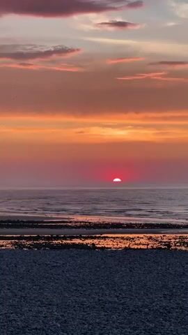 A time-lapse video captures a sunset over the ocean. The sun, a vibrant red orb, sinks below the horizon, casting a warm orange and pink glow across the sky and water. Clouds drift lazily overhead, tinged with purple and pink hues. The foreground shows a beach with dark pebbles and a body of water receding, revealing wet sand and rocks. In the distance, a small lighthouse stands on a jetty. A few birds can be seen in the sky at the beginning of the video, and a small group of people are visible on the beach towards the end.