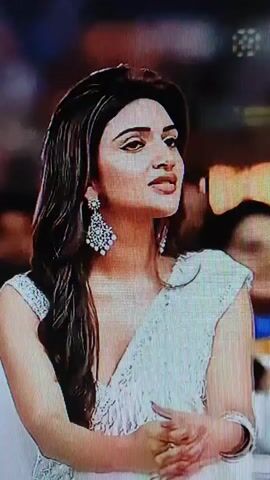 The video shows a series of close-up shots of individuals. The first shot is of a woman with long dark hair, wearing a white sari and large bejeweled earrings. The second shot shows a woman smiling broadly, with her hair partially pulled back, wearing a decorative top and a pink shawl. The third shot is a slightly blurry image of the same woman from the second shot, with another person partially visible behind her. The final shot is a close-up of a man with a beard, wearing a light-colored shirt, with a portion of a poster visible in the background. The video has a glitchy, digital aesthetic.