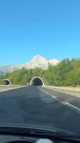 The video begins with a view from inside a vehicle, looking forward. The sky is clear and blue, with a large, rugged mountain peak visible in the distance. In the foreground, there is a two-lane asphalt road, leading towards two tunnels. The tunnel on the right is closer and more prominent, with a wide, arched entrance. The tunnel on the left is further away and partially obscured by trees. Lush green trees line the sides of the road and surround the tunnel entrances. The vehicle appears to be moving forward, approaching the right tunnel. As the video progresses, the vehicle enters the tunnel. The interior of the tunnel is dimly lit, with lights visible along the ceiling. The road continues forward, with white lines marking the lanes. The view from inside the car, including the dashboard and windshield wipers, is visible at the bottom of the frame. The tunnel is constructed of concrete, with visible seams and structural elements. The video shows the vehicle traveling through the tunnel, with the entrance receding and the darkness ahead continuing.
