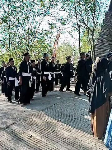 A group of people dressed in black martial arts uniforms with white sashes are walking in a procession outdoors. They are on a paved path, with trees and foliage in the background. There are flags with writing on them visible. Some of the individuals are wearing hats. The overall atmosphere appears to be ceremonial or a parade.