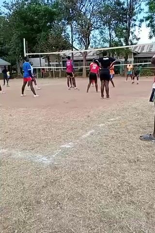 The video shows a volleyball match in progress on a grassy, dirt court. Several players in colorful jerseys and shorts are positioned on both sides of a net. A volleyball is visible on the ground, having just been played. Spectators stand along the edges of the court. A building and trees are visible in the background. One man wearing a red shirt and blue shorts stands in the foreground, and then another man wearing a white shirt with black details and blue shorts walks across the foreground, in front of the camera.