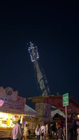 A nighttime amusement park scene features a tall, illuminated ride with lights changing colors from purple to green. The ride is angled upwards against a dark sky. In the foreground, a food stall with bright lights and signage is visible, along with a crowd of people standing and walking. An "Ausgang Exit" sign is mounted on a pole. The atmosphere suggests a lively and festive environment.